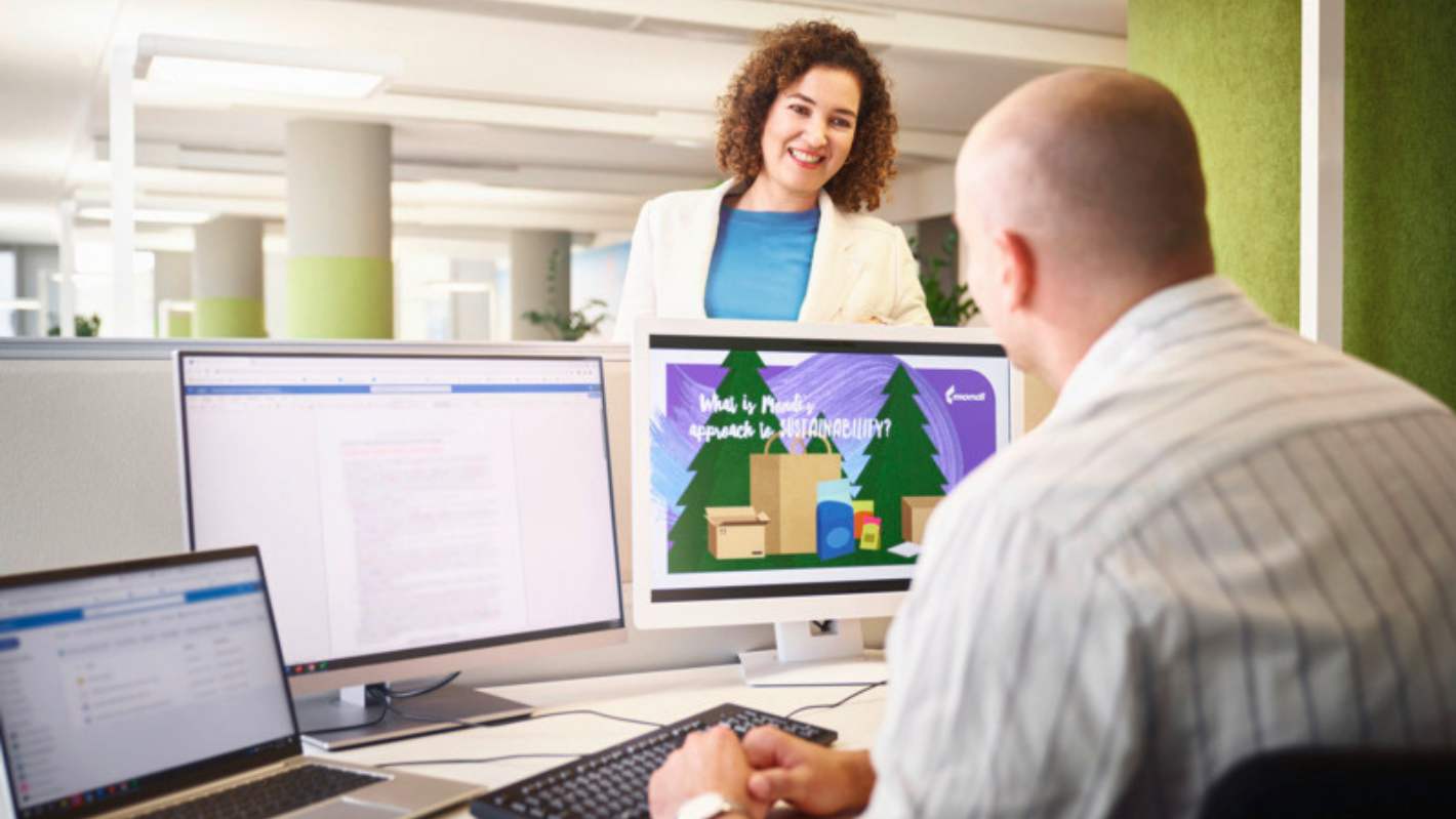 2 Mondi colleagues in an office, a man visible from the back in front of 3 screens, talking to a woman who is opposite of him and looking at him in a friendly manner